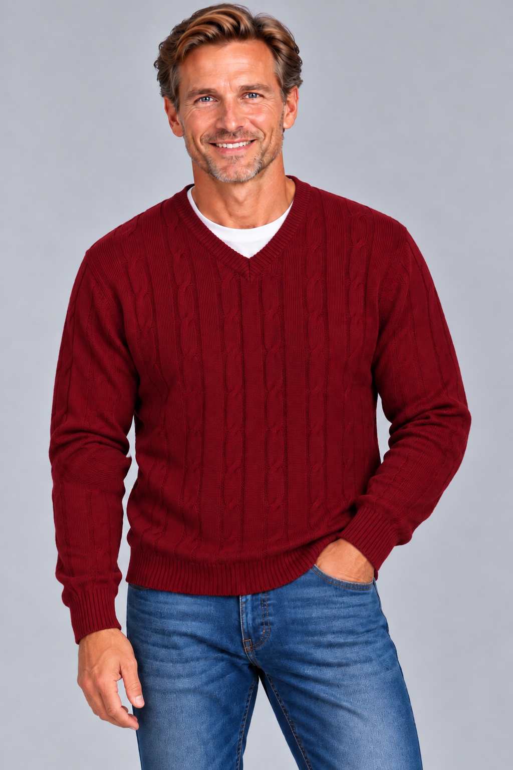 A smiling man wearing a burgundy cable-knit V-neck sweater over a white undershirt and blue jeans, standing against a solid gray studio background.