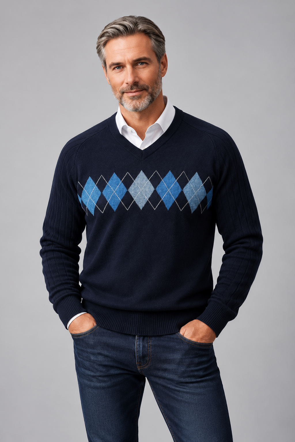 A man wearing a navy blue v-neck sweater with an argyle diamond pattern in shades of blue and gray, layered over a white collared shirt and paired with dark blue jeans, against a solid gray studio background.