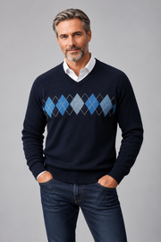 A man wearing a navy blue v-neck sweater with an argyle diamond pattern in shades of blue and gray, layered over a white collared shirt and paired with dark blue jeans, against a solid gray studio background.