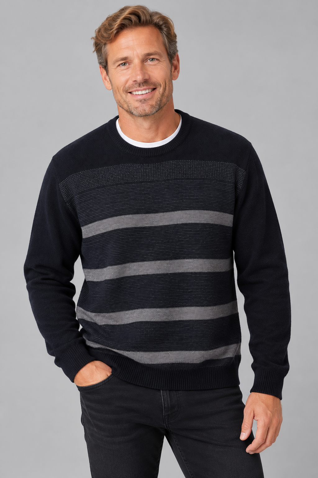 A smiling man wearing a dark navy crewneck sweater with horizontal gray stripes, paired with dark jeans, against a solid gray studio background.