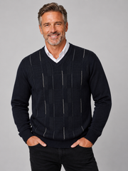 A smiling middle-aged man wearing a dark navy blue V-neck sweater with vertical textured stripe panels over a white collared shirt, paired with black pants, against a solid gray studio background.