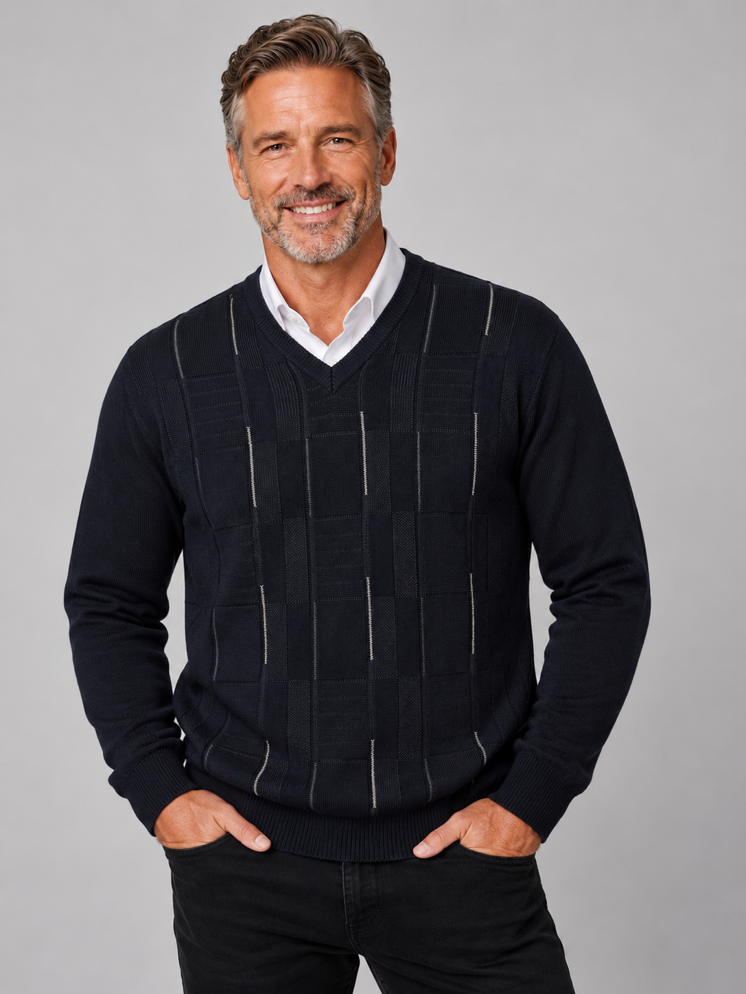 A smiling middle-aged man wearing a dark navy blue V-neck sweater with vertical textured stripe panels over a white collared shirt, paired with black pants, against a solid gray studio background.