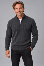 A smiling man wearing a dark charcoal gray ribbed quarter-zip sweater over a white undershirt and black pants, standing against a solid gray studio background.