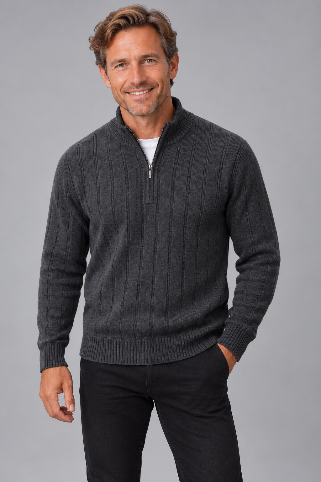 A smiling man wearing a dark charcoal gray ribbed quarter-zip sweater over a white undershirt and black pants, standing against a solid gray studio background.