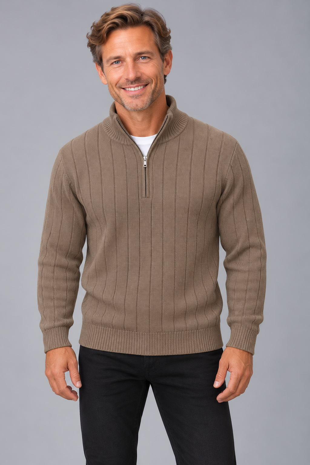 A smiling man wearing a taupe ribbed quarter-zip sweater over a white undershirt and black pants, standing against a solid gray studio background.