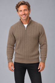 A smiling man wearing a taupe ribbed quarter-zip sweater over a white undershirt and black pants, standing against a solid gray studio background.