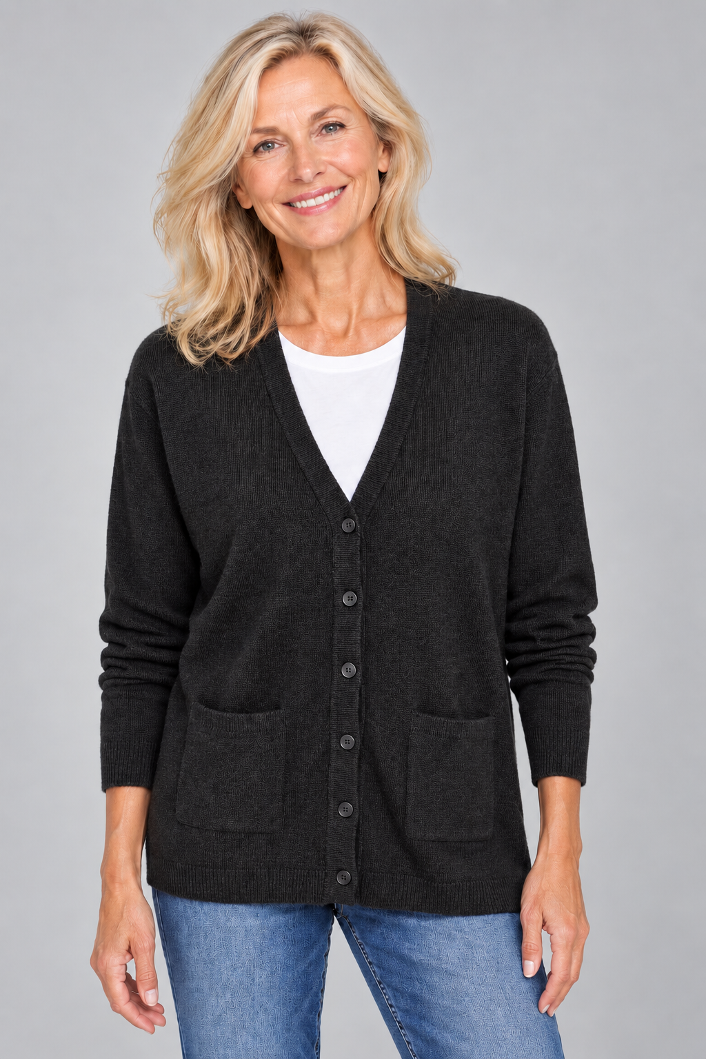 A smiling blonde woman wearing a dark charcoal gray button-up cardigan over a white t-shirt and blue jeans, photographed against a solid gray studio background.
