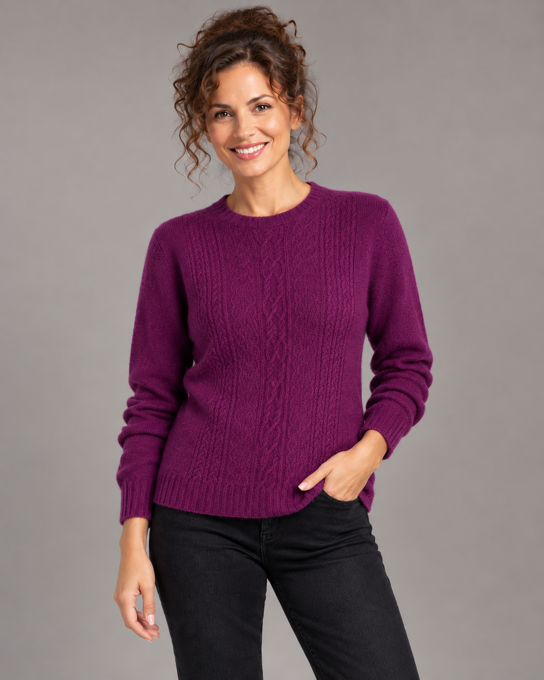 A woman wearing a purple cable-knit crewneck sweater and black pants, standing against a solid gray studio background.