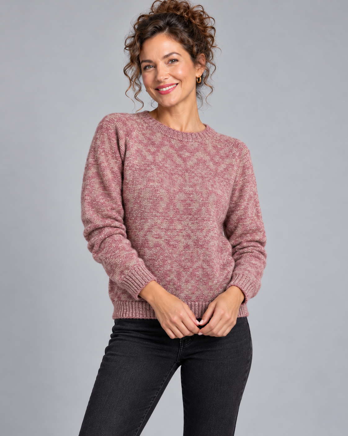 A woman wearing a dusty rose and beige marled knit crewneck sweater with black jeans, standing against a solid gray studio background.