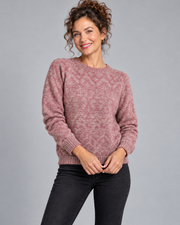 A woman wearing a dusty rose and beige marled knit crewneck sweater with black jeans, standing against a solid gray studio background.