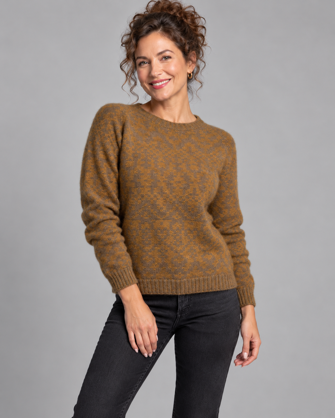 A woman modeling a brown and gold patterned knit sweater with black jeans, standing against a solid gray studio background.