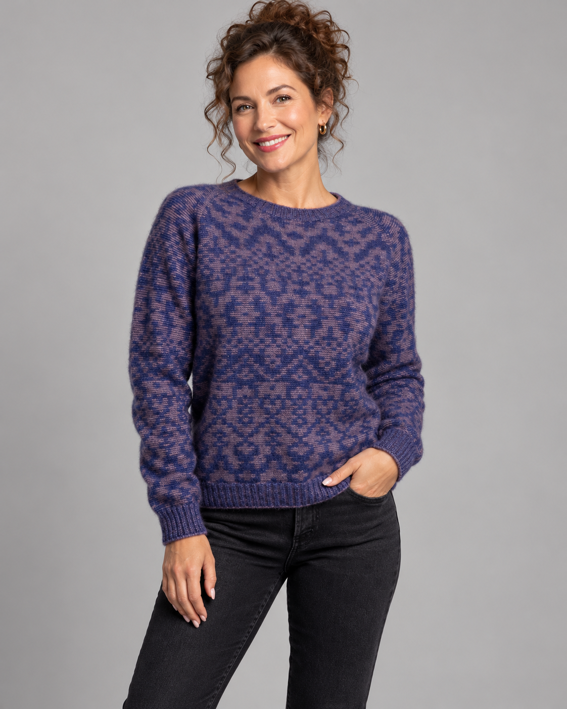 A woman wearing a purple and blue patterned knit sweater with black jeans, standing against a solid gray studio background.