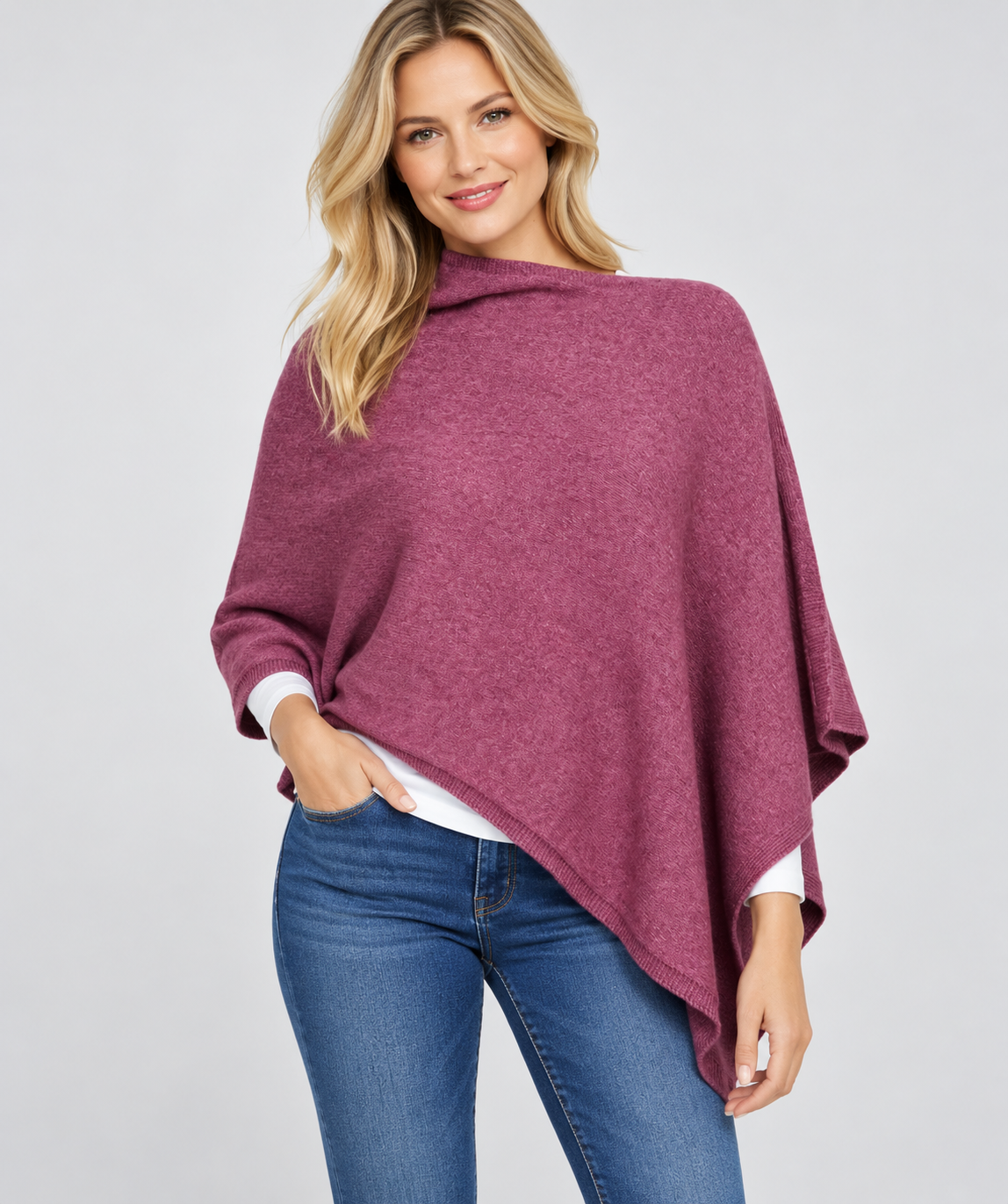 A woman modeling a heathered dusty rose purple poncho over a white long-sleeve shirt and blue jeans, photographed against a solid light gray studio background.