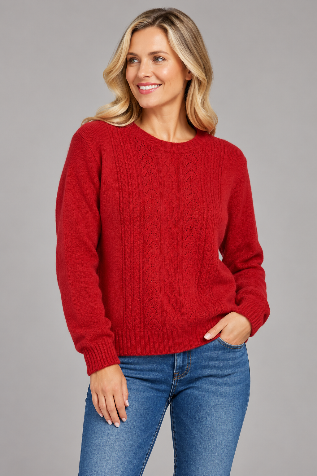 A woman wearing a red cable-knit crewneck sweater and blue jeans, standing against a solid gray studio background.